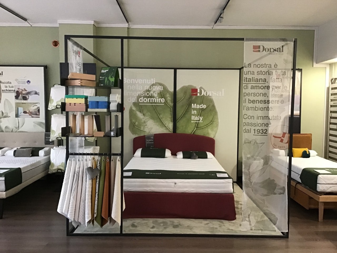 Letto matrimoniale con testata rossa in esposizione, tessuti e campioni, sfondo con foglia e testo pubblicitario Dorsal Made…