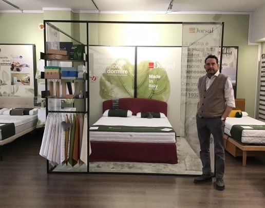 Letto matrimoniale con testiera rossa, materasso bianco con rifiniture verdi, uomo con gilet grigio accanto, scaffale con…