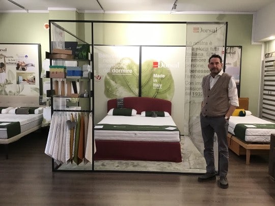 Letto matrimoniale con testiera rossa, materasso bianco con rifiniture verdi, uomo con gilet grigio accanto, scaffale con…