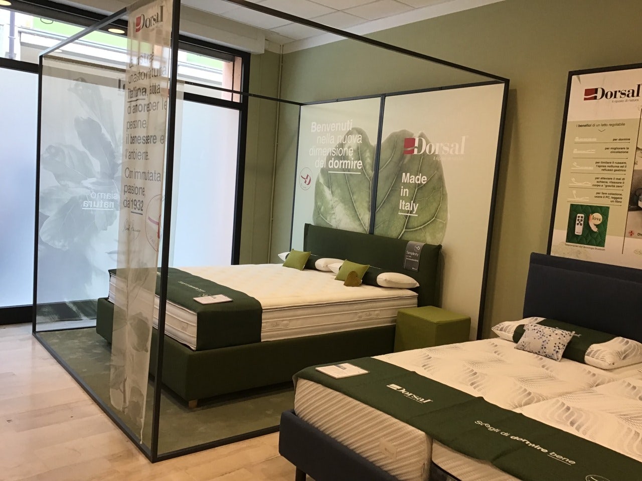 Due letti matrimoniali con testiere imbottite in esposizione in negozio, con dettagli verdi e cartelli promozionali Dorsal…