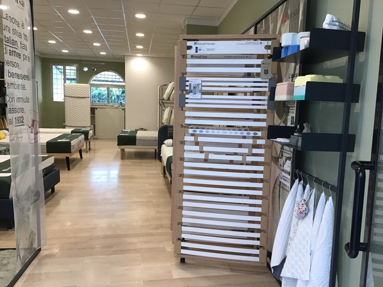 Negozio con esposizione di materassi, reti e biancheria da letto su pavimento in legno chiaro e pareti verdi.