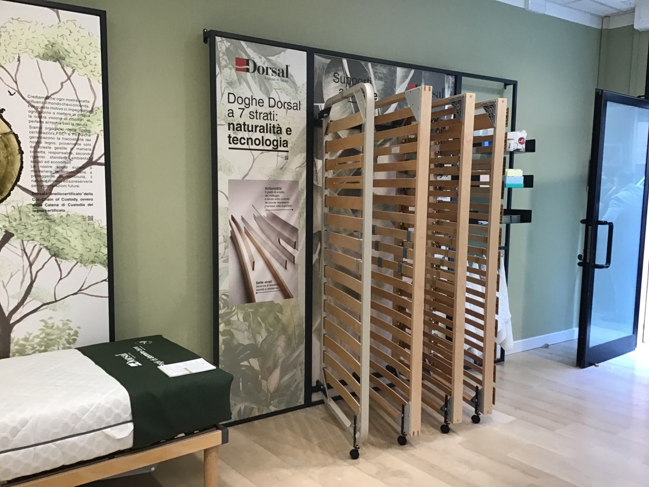 Lettiere a doghe di legno su ruote esposte in showroom con pareti verdi e poster promozionali sulla sostenibilità del legno.
