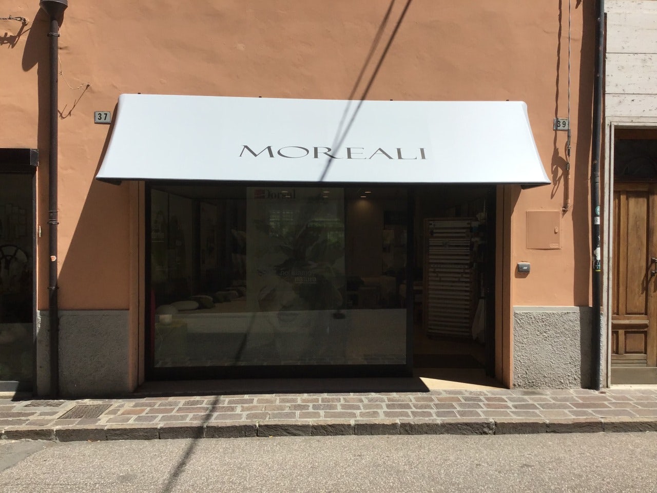 Vetrina di negozio con tenda bianca e scritta Moreali su muro arancione e marciapiede in pietra davanti.
