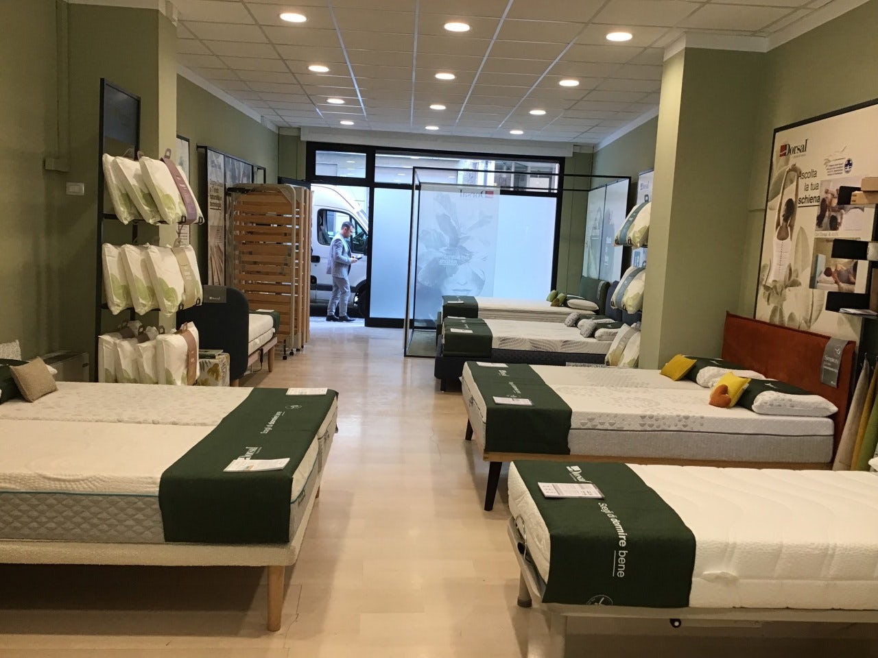 Negozio di materassi con diversi modelli esposti in fila e scaffali con cuscini, pavimento in legno chiaro.
