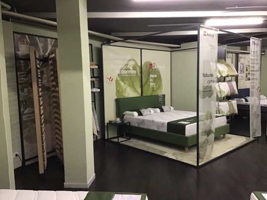 Zona espositiva con letto verde, cuscini bianchi, pareti vetrate con scritte Benvenuti nella nuova dimensione del dormire e…