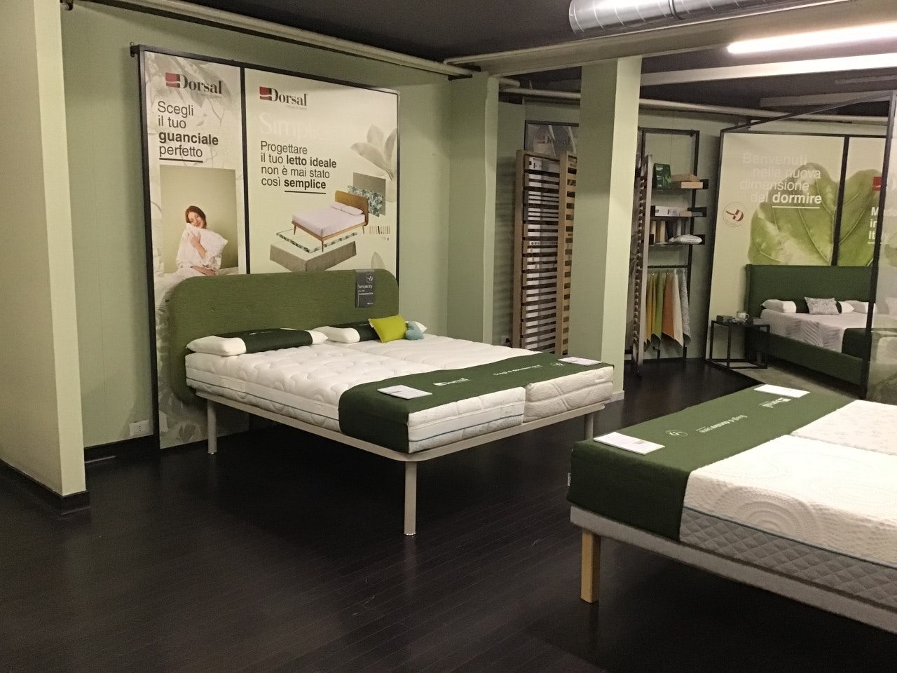 Letti matrimoniali con testiera verde e materassi bianchi in esposizione su pavimento scuro, sfondo con poster pubblicitari…