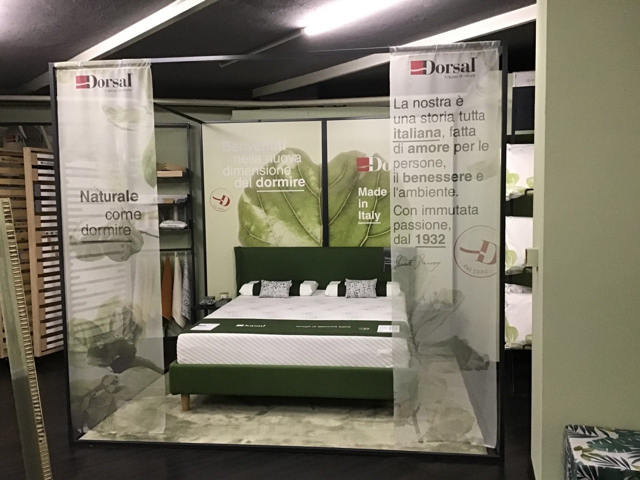 Letto con testata verde e materasso bianco, due tende trasparenti con scritte promozionali Dorsal e sfondo con foglia verde.