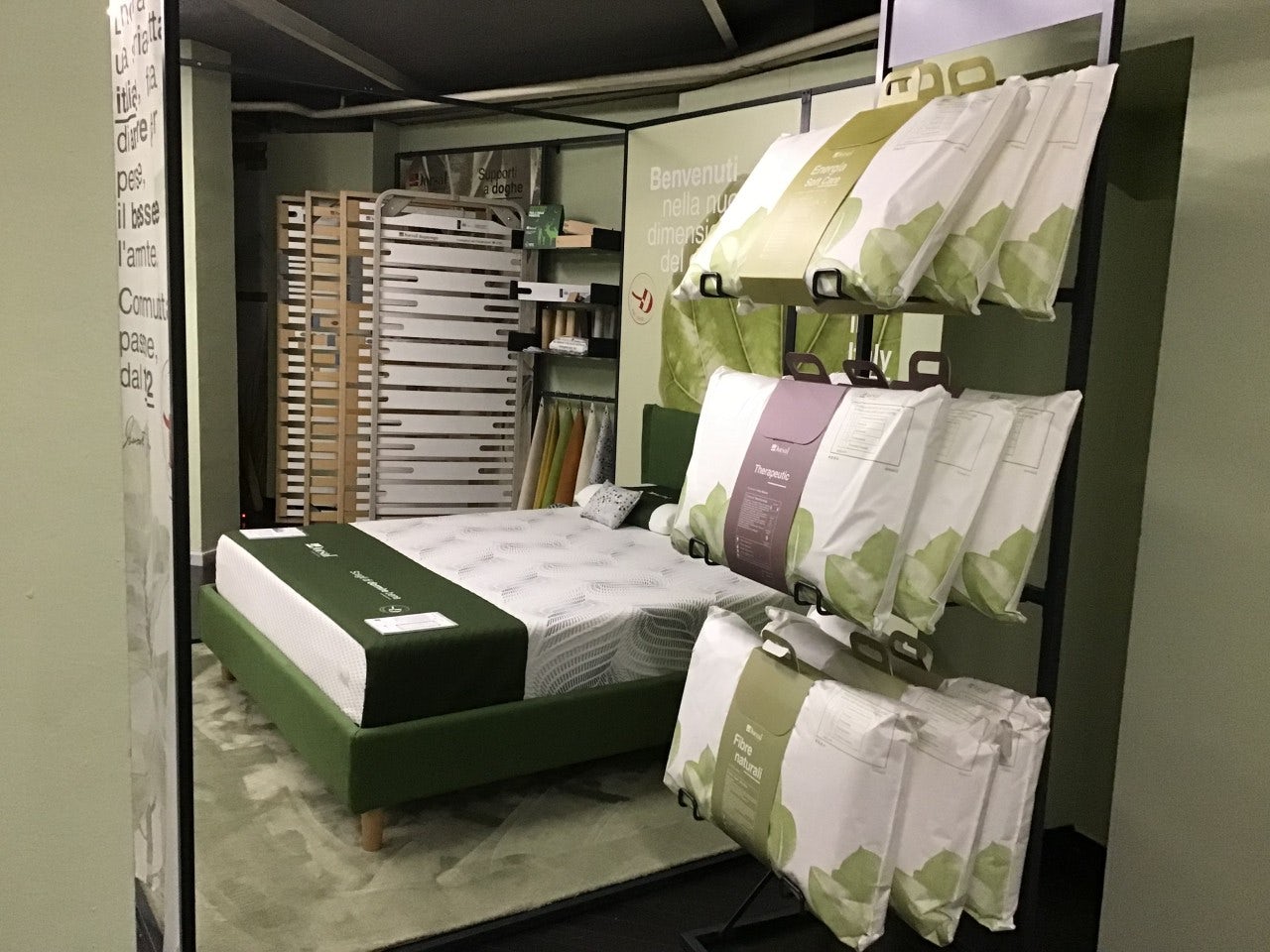Letto con struttura verde, materasso bianco e pacchi di biancheria per letto appesi su supporto metallico nero.