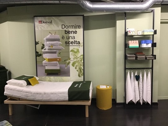 Letto singolo con materasso e cuscini bianchi su struttura in legno, poster con testo promozionale e scaffale nero con…
