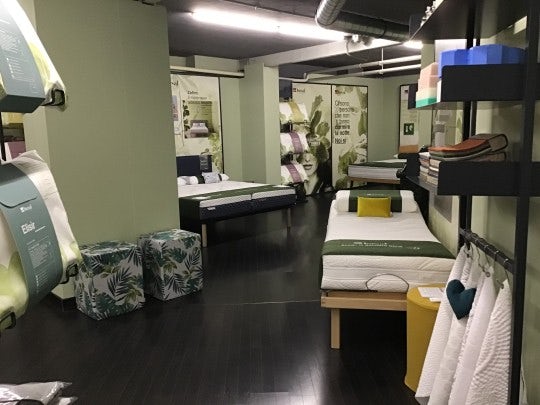 Negozio con letti esposti, biancheria da letto, cuscini decorativi, due pouf con motivo foglie verdi e pavimento scuro.