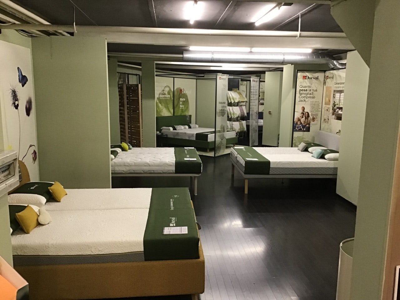 Showroom con diversi letti singoli e matrimoniali con copriletto verde e pareti color salvia con decorazioni floreali.