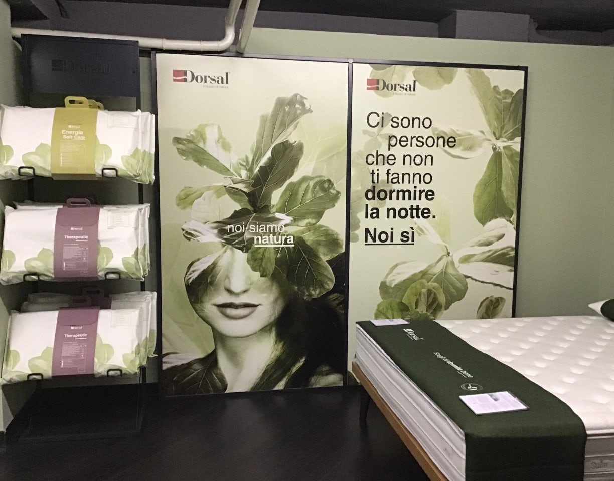 Letto con materasso bianco e verde, cuscini su ripiano nero, poster con volto femminile e scritta Noi siamo natura e Dormire…