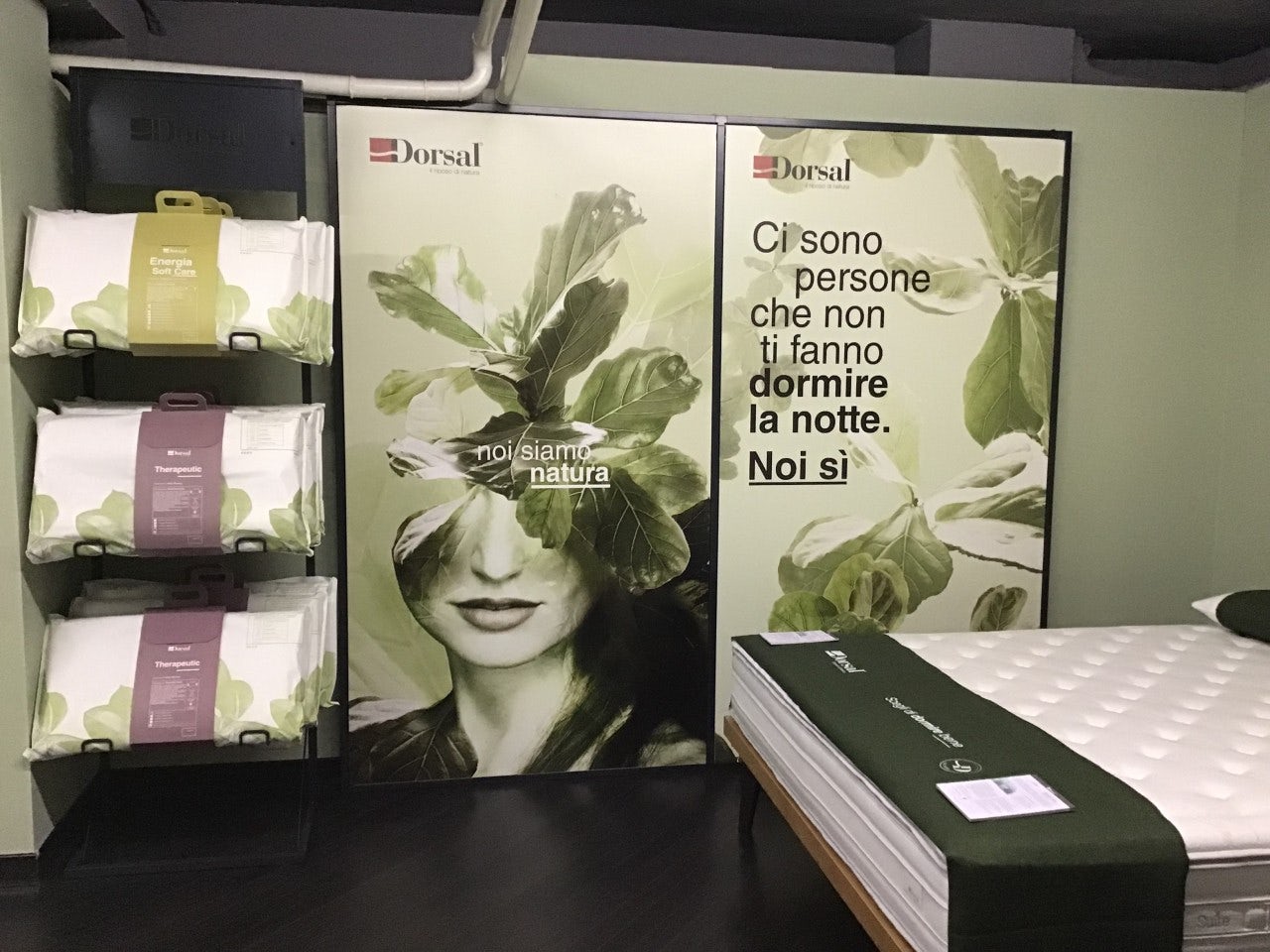 Letto con materasso bianco e verde, cuscini su ripiano nero, poster con volto femminile e scritta Noi siamo natura e Dormire…