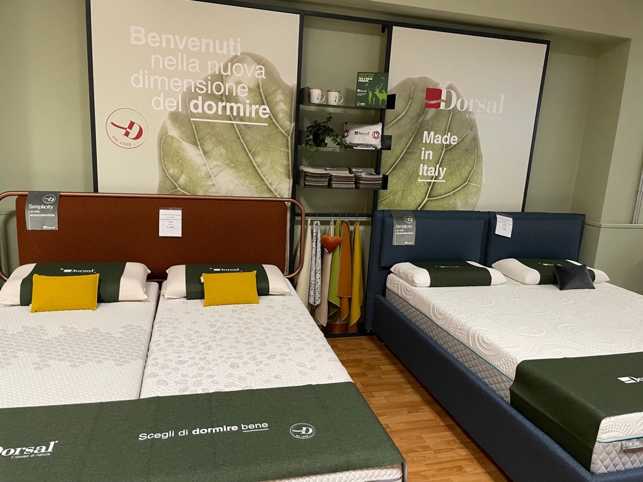 Due letti matrimoniali con testiere imbottite, cuscini gialli e posizione in showroom con scritte promozionali sullo sfondo.