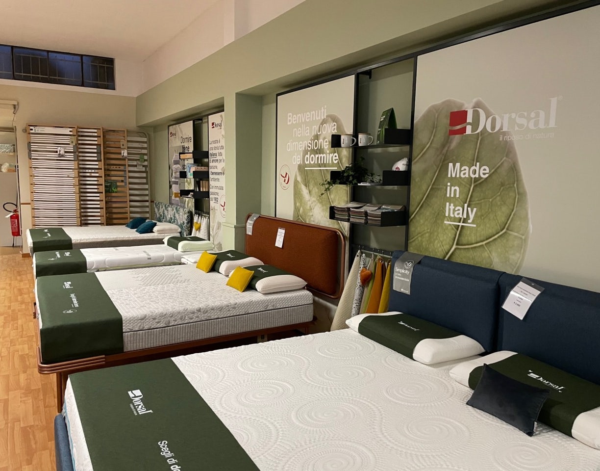 Negozio con diversi letti e materassi con coperture verdi, testiere imbottite e manifesto con scritte Benvenuti nella nuova…