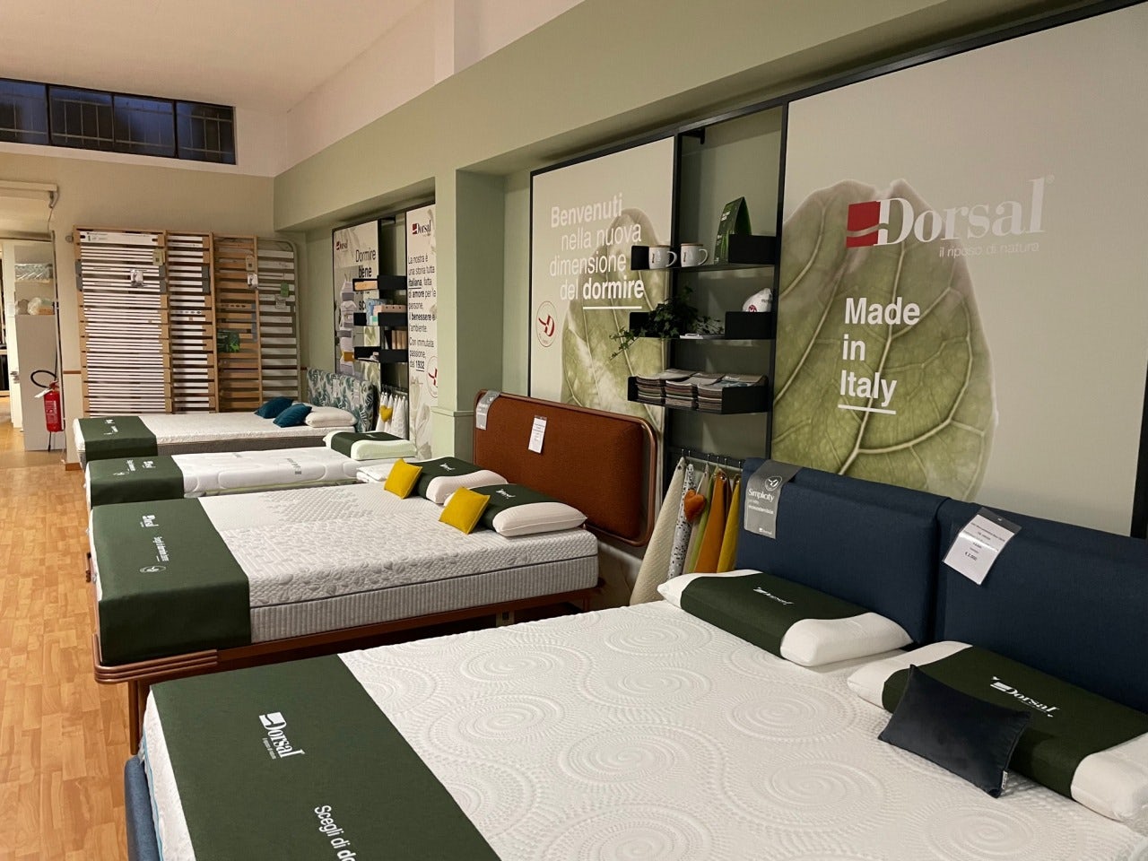 Negozio con diversi letti e materassi con coperture verdi, testiere imbottite e manifesto con scritte Benvenuti nella nuova…