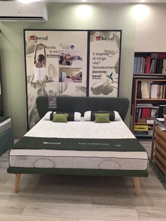Letto con materasso Dorsal e cuscini, testata verde e pannelli pubblicitari su parete con messaggi su salute della schiena.