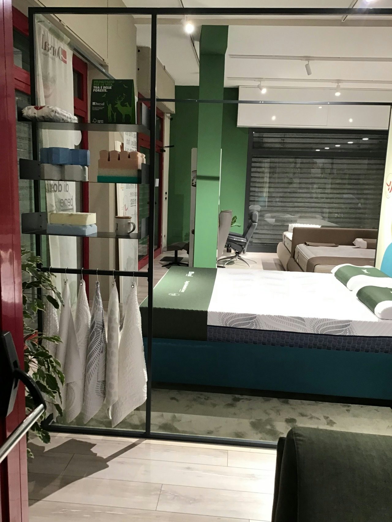 Esposizione di materassi con vari cuscini e asciugamani bianchi appesi in negozio di arredamento.
