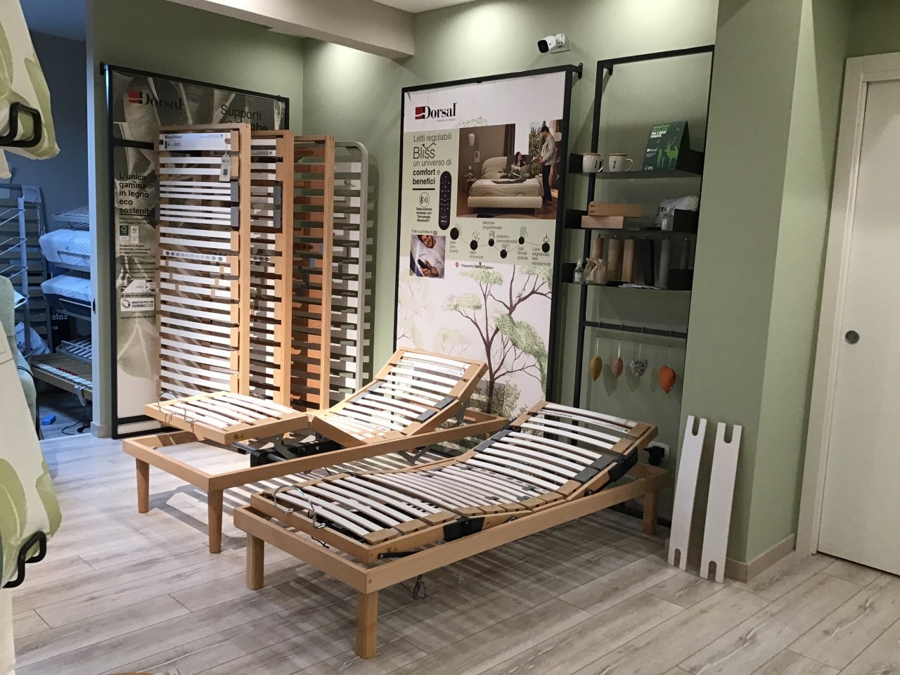 Due letti regolabili in legno con doghe esposte in showroom con poster promozionali e scaffalature verdi.