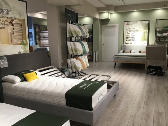 Negozio con letti e materassi esposti, pareti chiare e pavimento in legno chiaro, cuscini e coperte in verde scuro.
