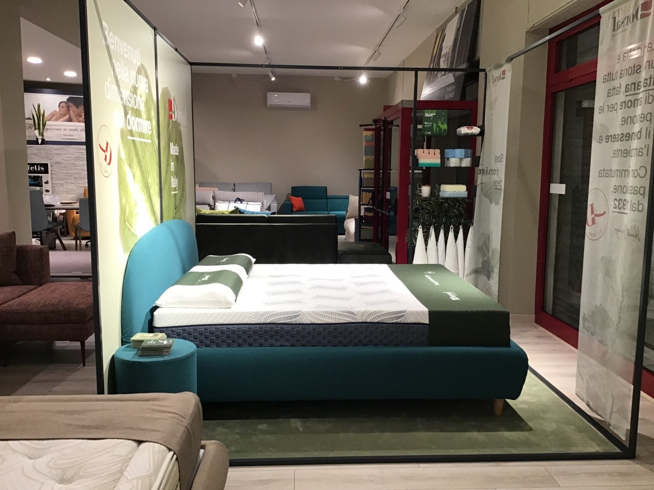 Letto matrimoniale con testiera e complemento turchese, materasso bianco con cuscini verdi esposto in showroom arredamento.