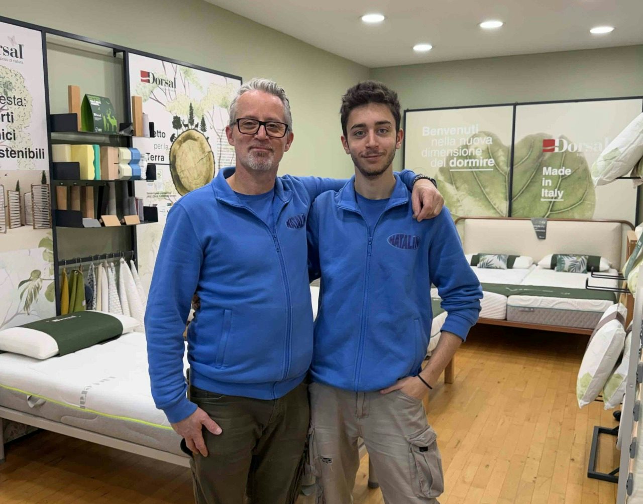 Due uomini con felpe blu posano in negozio di letti e materassi con pavimento in legno e segni Dorsal Made in Italy.