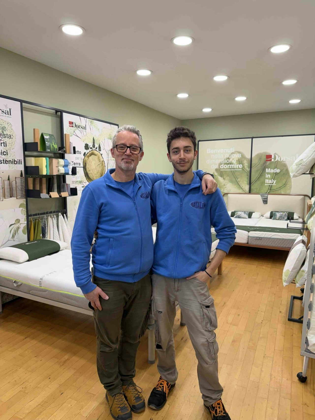 Due uomini con felpe blu posano in negozio di letti e materassi con pavimento in legno e segni Dorsal Made in Italy.