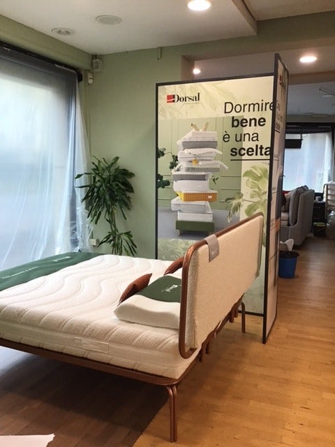 Letto con materasso bianco e biancheria verde in stanza con pavimento in legno e pianta vicino a grande finestra velata.