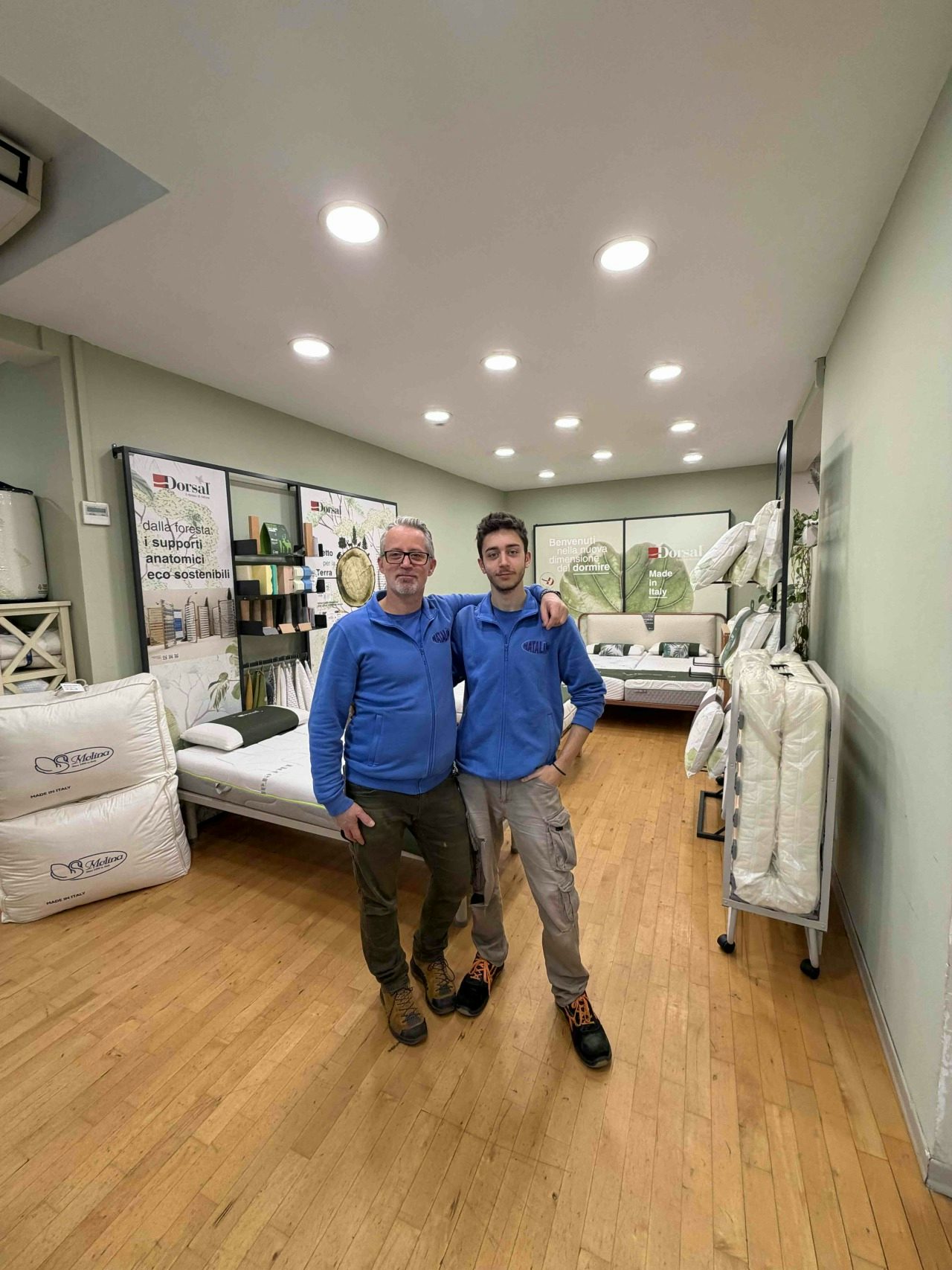 Due uomini con felpe blu in un negozio di materassi con parquet e illuminazione a soffitto intensa
