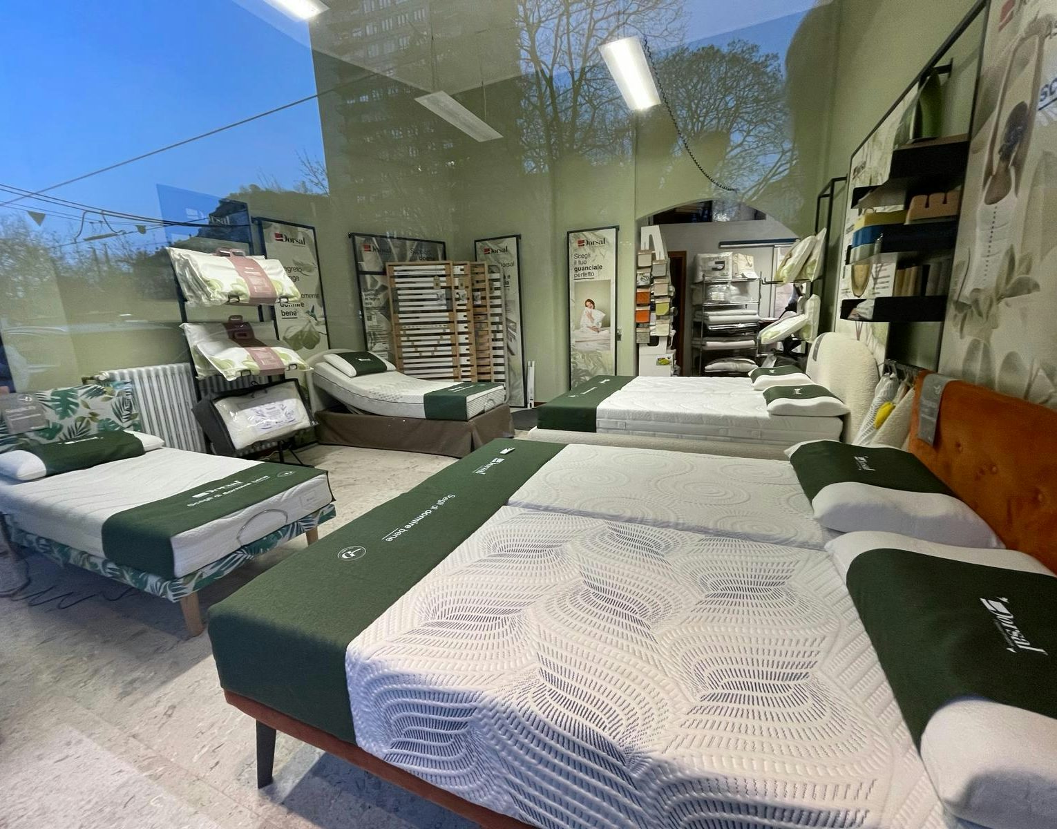 Negozio con letti singoli e matrimoniali con coprimaterassi verdi e cuscini, scaffali con biancheria e poster pubblicitari…