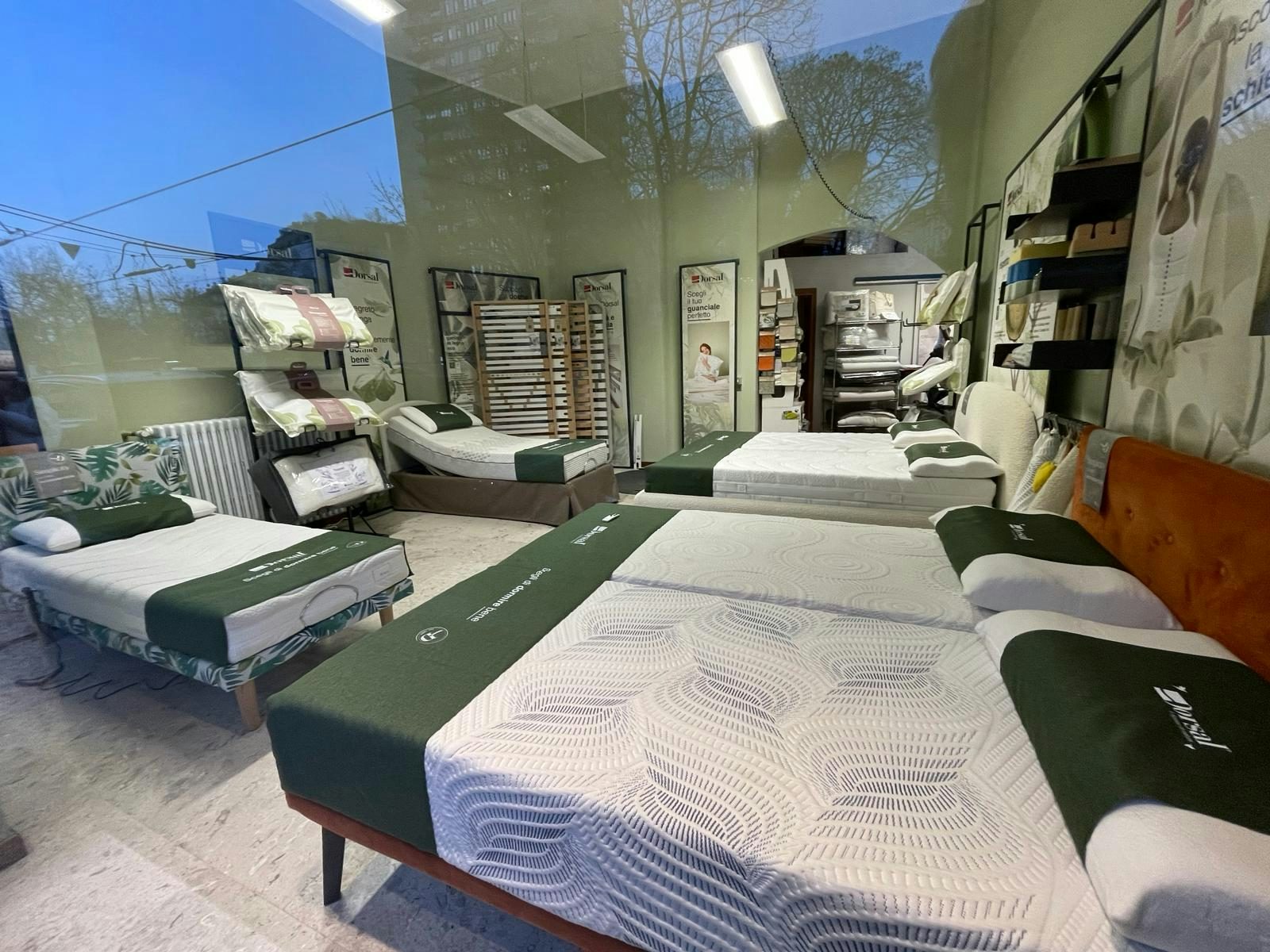 Negozio con letti singoli e matrimoniali con coprimaterassi verdi e cuscini, scaffali con biancheria e poster pubblicitari…
