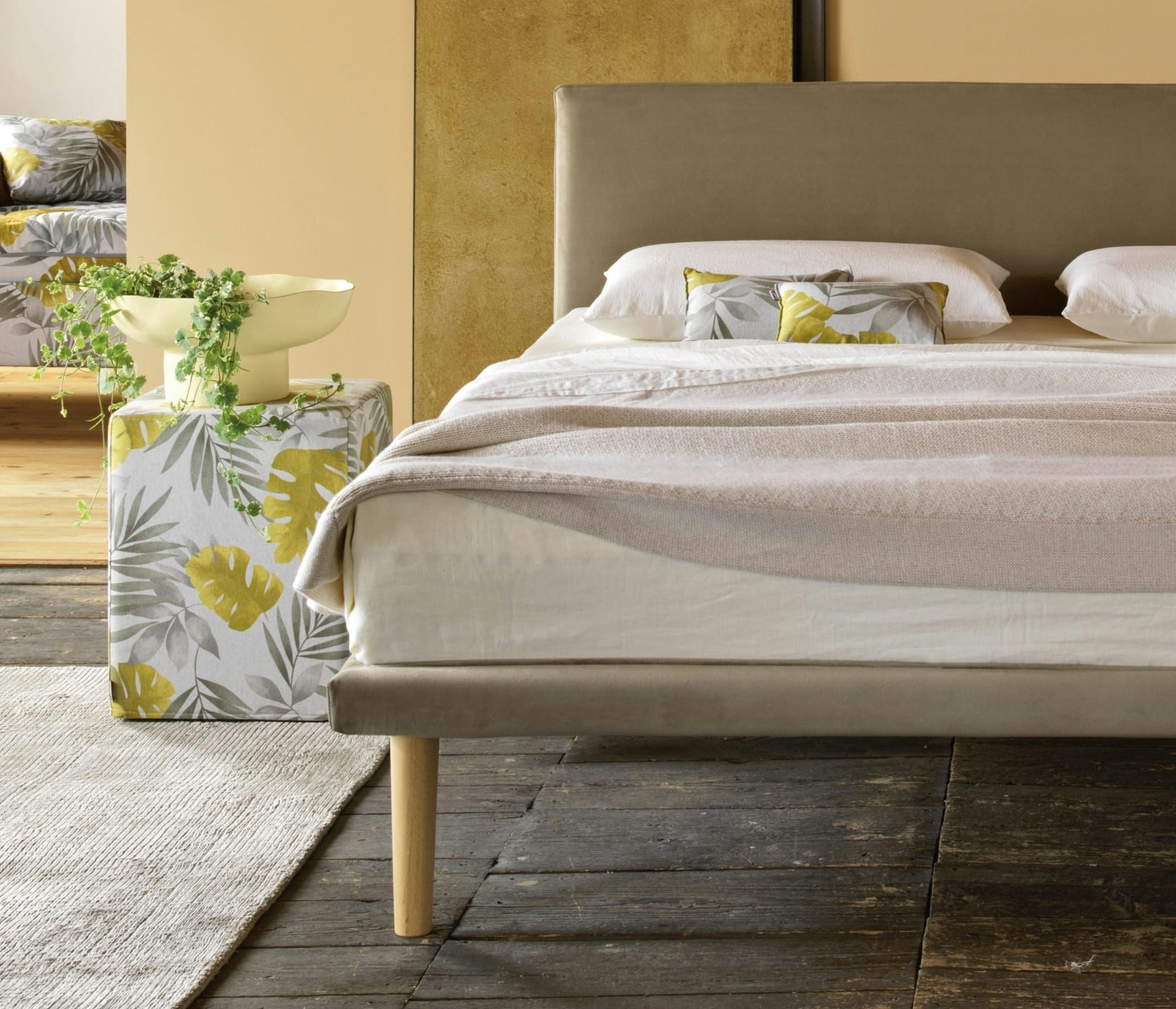 Letto beige con piedi in legno chiaro, cuscini decorativi con motivo foglie verdi e gialle, tavolino rivestito e vaso con…