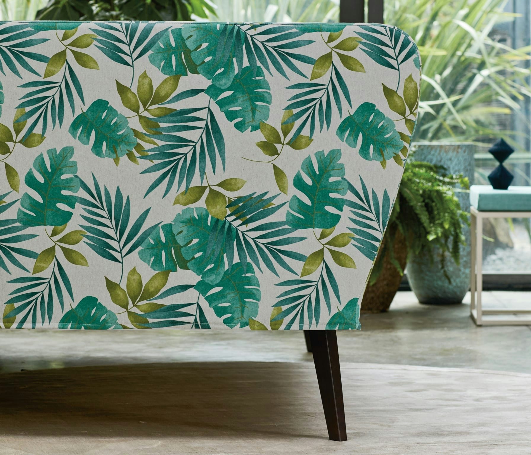 Parte laterale di divano con tessuto fantasia foglie tropicali su sfondo chiaro, gambo in legno scuro, pavimento chiaro.