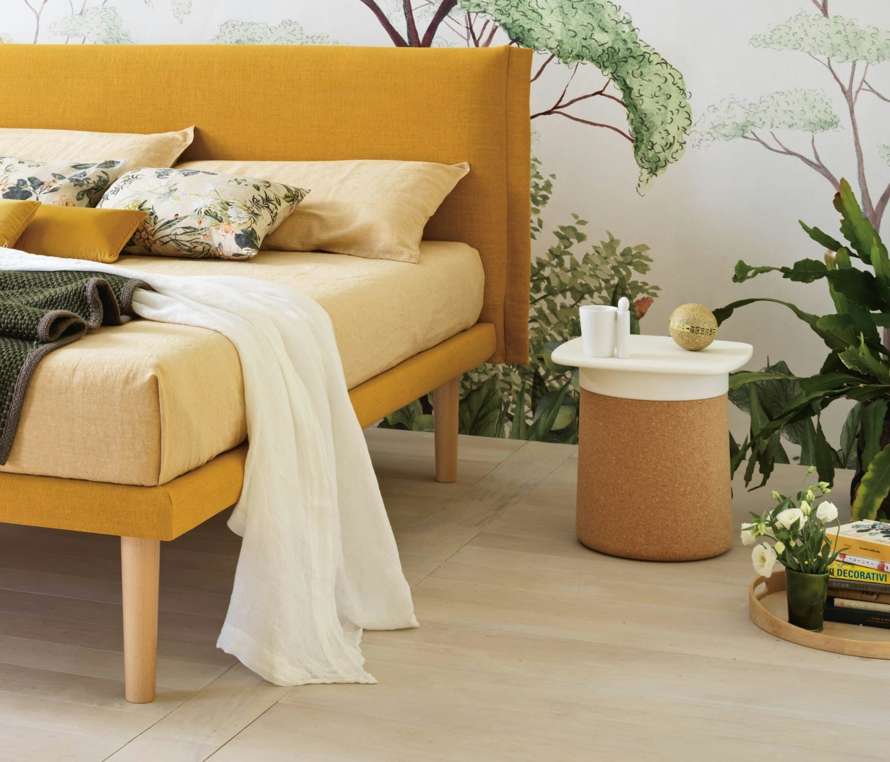 Letto con struttura gialla e cuscini floreali, accanto tavolino bianco e sughero con oggetti decorativi e piante verdi.