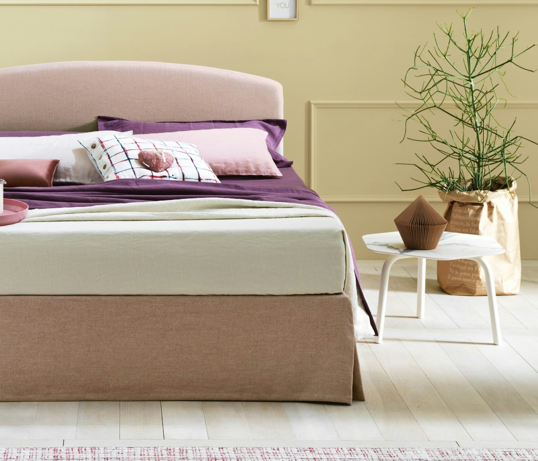 Letto beige con cuscini color pastello e viola, tavolino bianco con decorazione marrone, pianta in vaso marrone di carta.