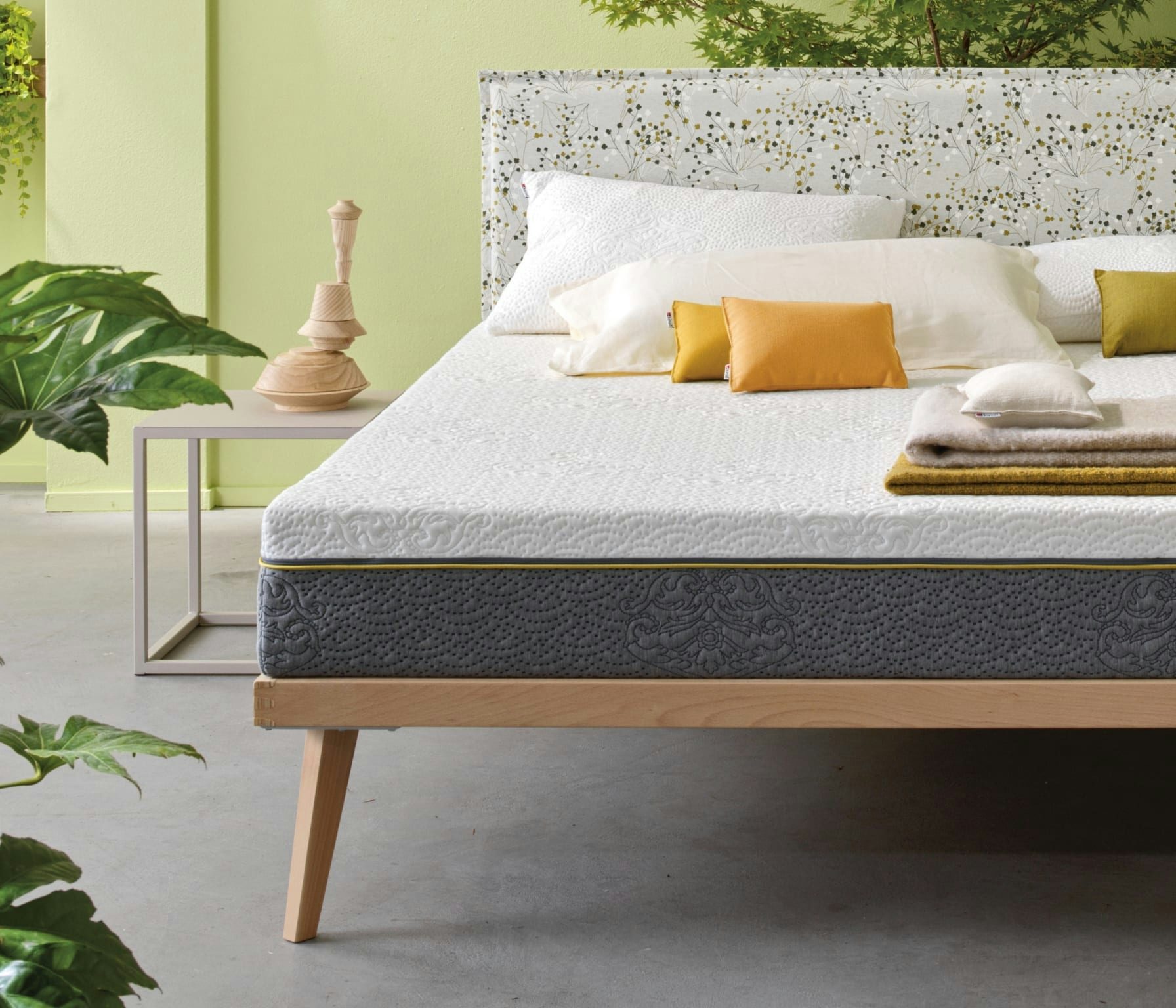 Letto con struttura in legno chiaro, materasso bianco con bordo grigio, cuscini gialli e bianchi, accanto tavolino e piante…