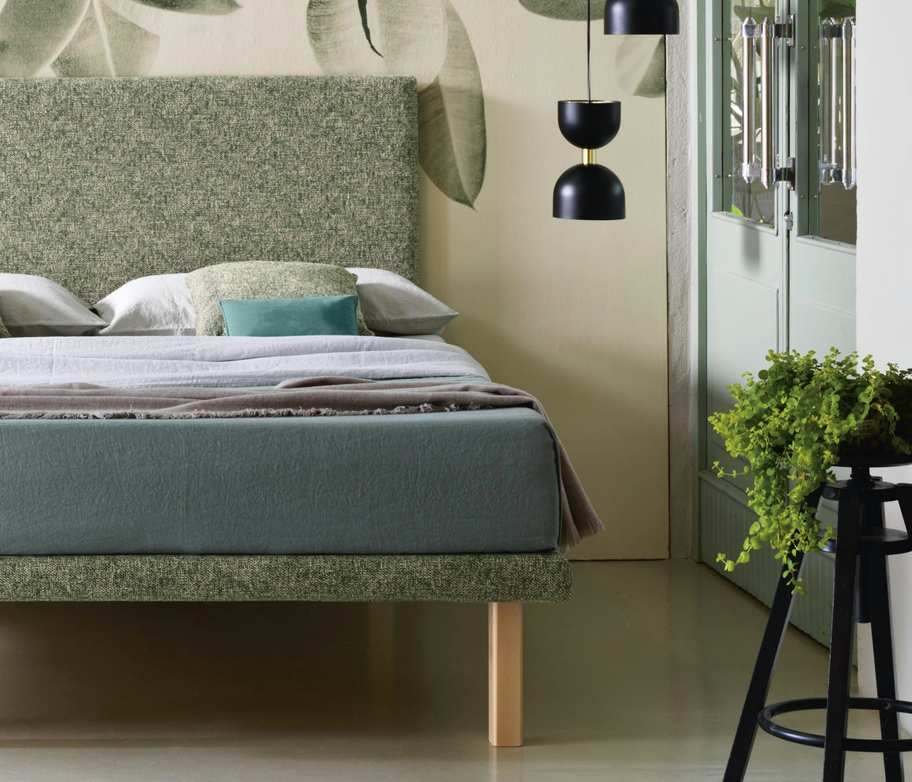 Letto matrimoniale verde con testiera imbottita, lenzuola e cuscini, lampada nero opaco e pianta su sgabello nero.