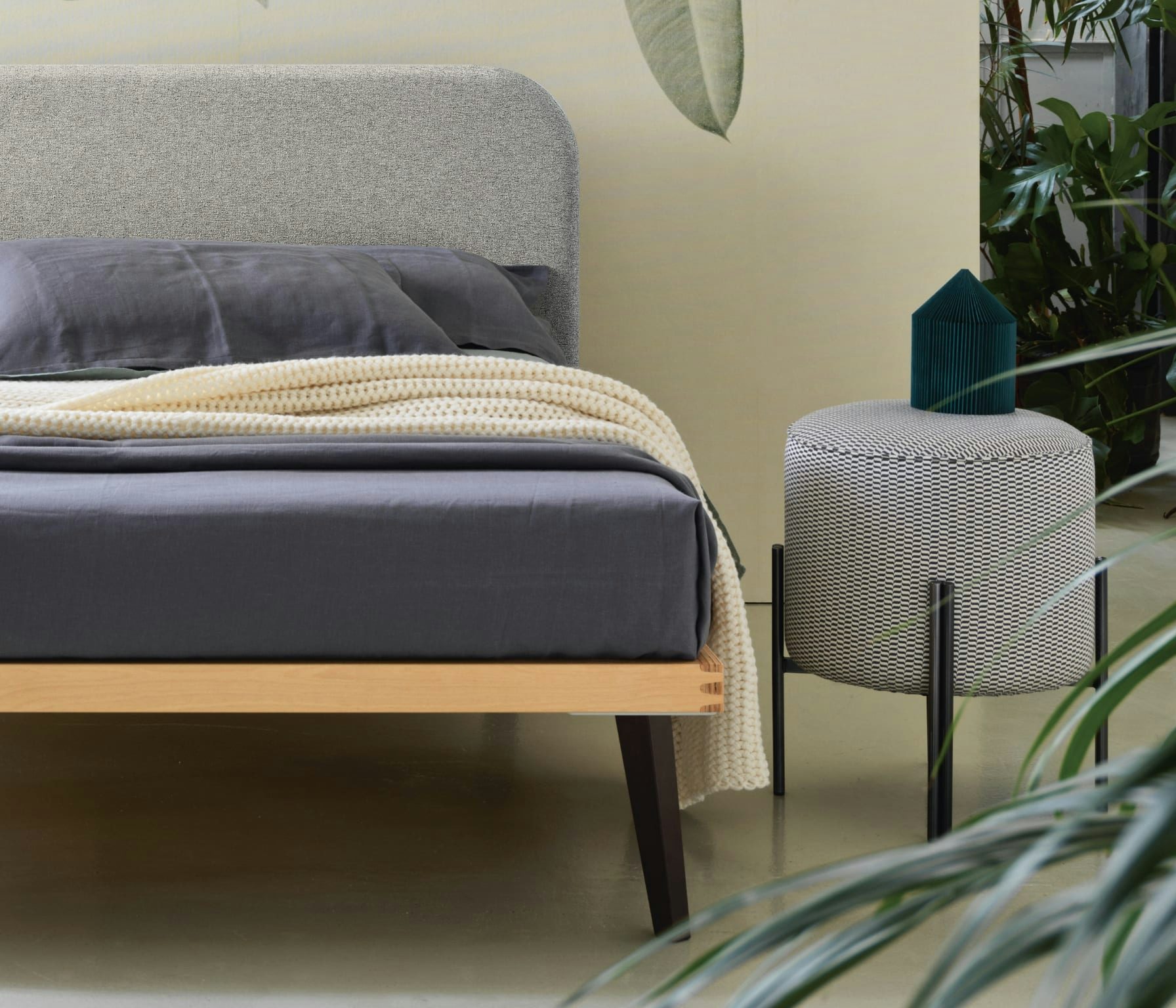 Letto grigio con copriletto blu scuro e coperta a maglia chiara, accanto pouf tessuto geometrico con vaso blu.