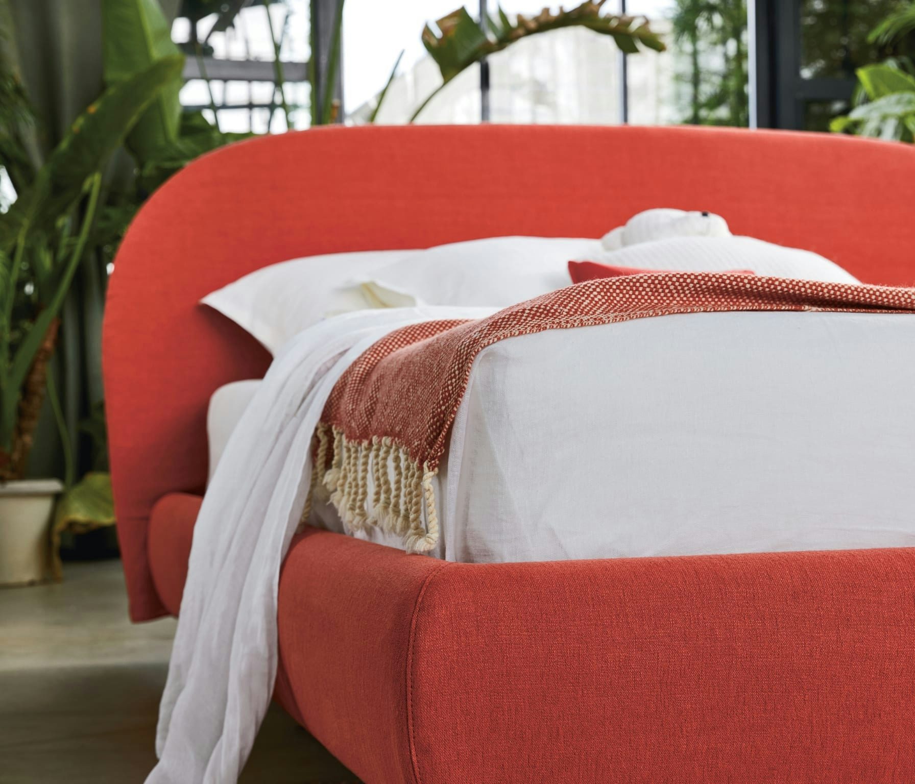 Letto matrimoniale con struttura imbottita rossa, coperta bianca e plaid arancione con frange in stanza con piante.