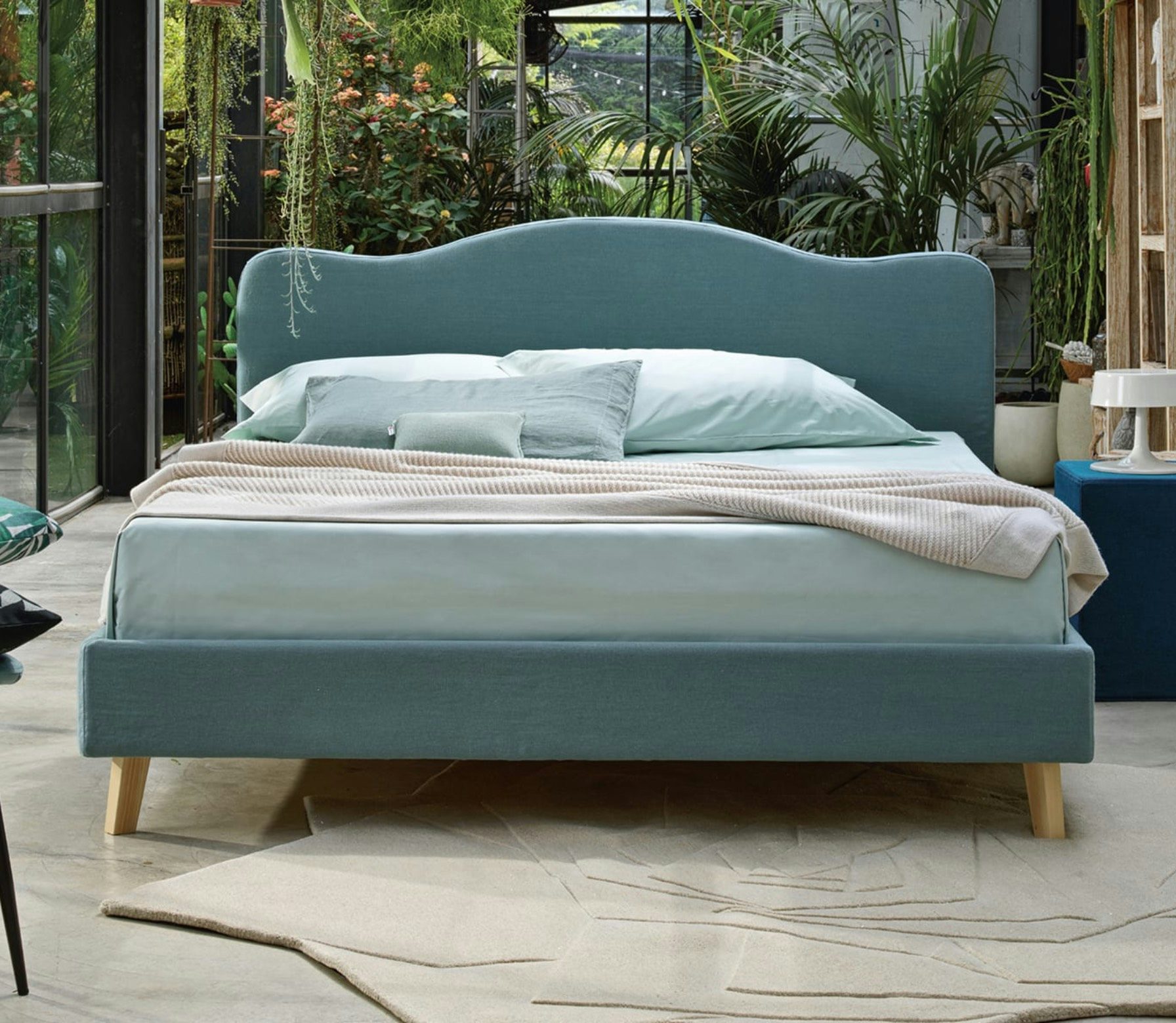 Letto matrimoniale con struttura e testiera imbottite azzurro polvere, coperte beige e cuscini turchesi, piante verdi sullo…