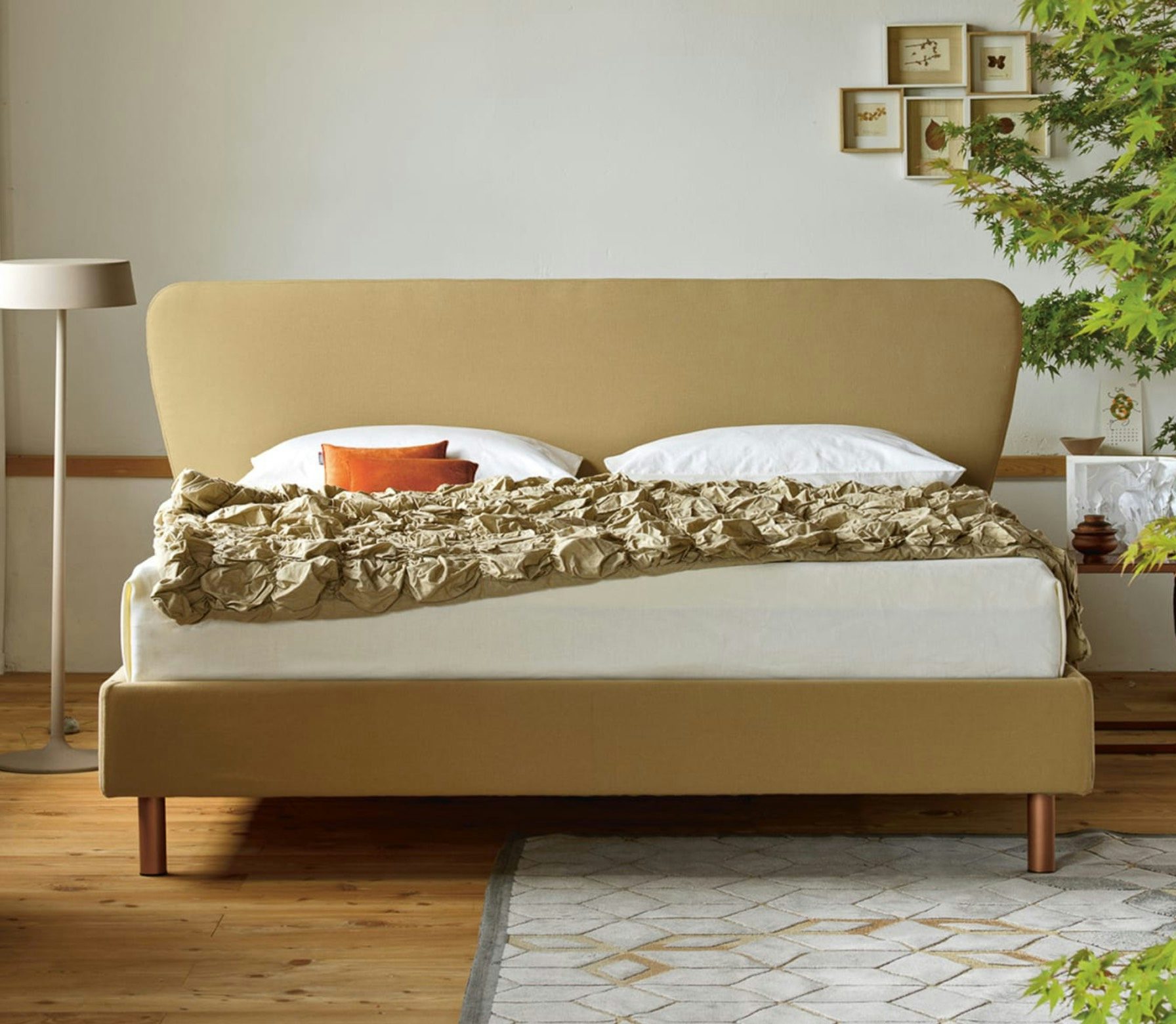 Letto matrimoniale con testiera e base beige, coperta arricciata color sabbia, cuscini bianchi e arancioni, lampada da…
