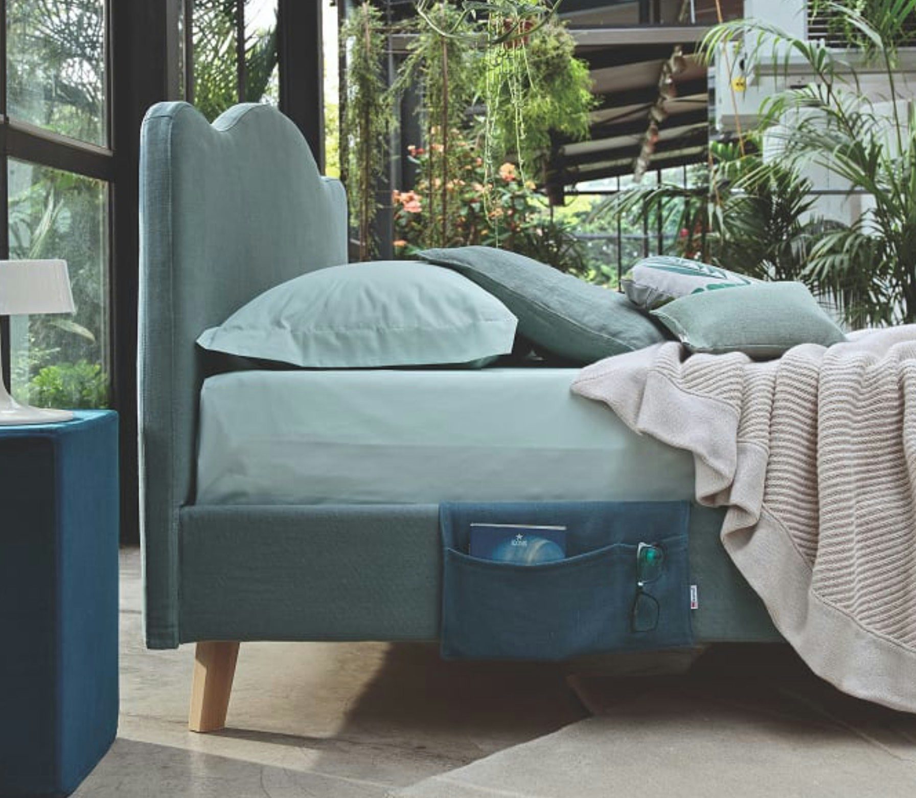 Letto tessuto verde acqua con testiera sagomata, cuscini, coperta beige e tasca laterale con libro e occhiali.