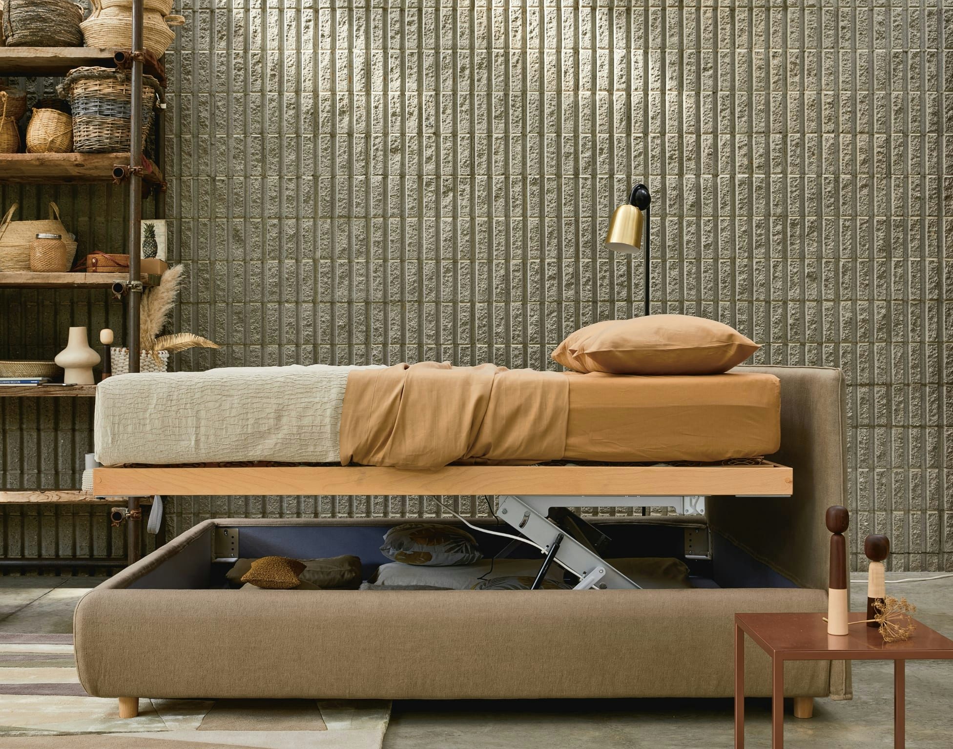 Letto contenitore beige con materasso sollevato, cuscino marrone, scaffale con cesti e parete grigia a mattoni.