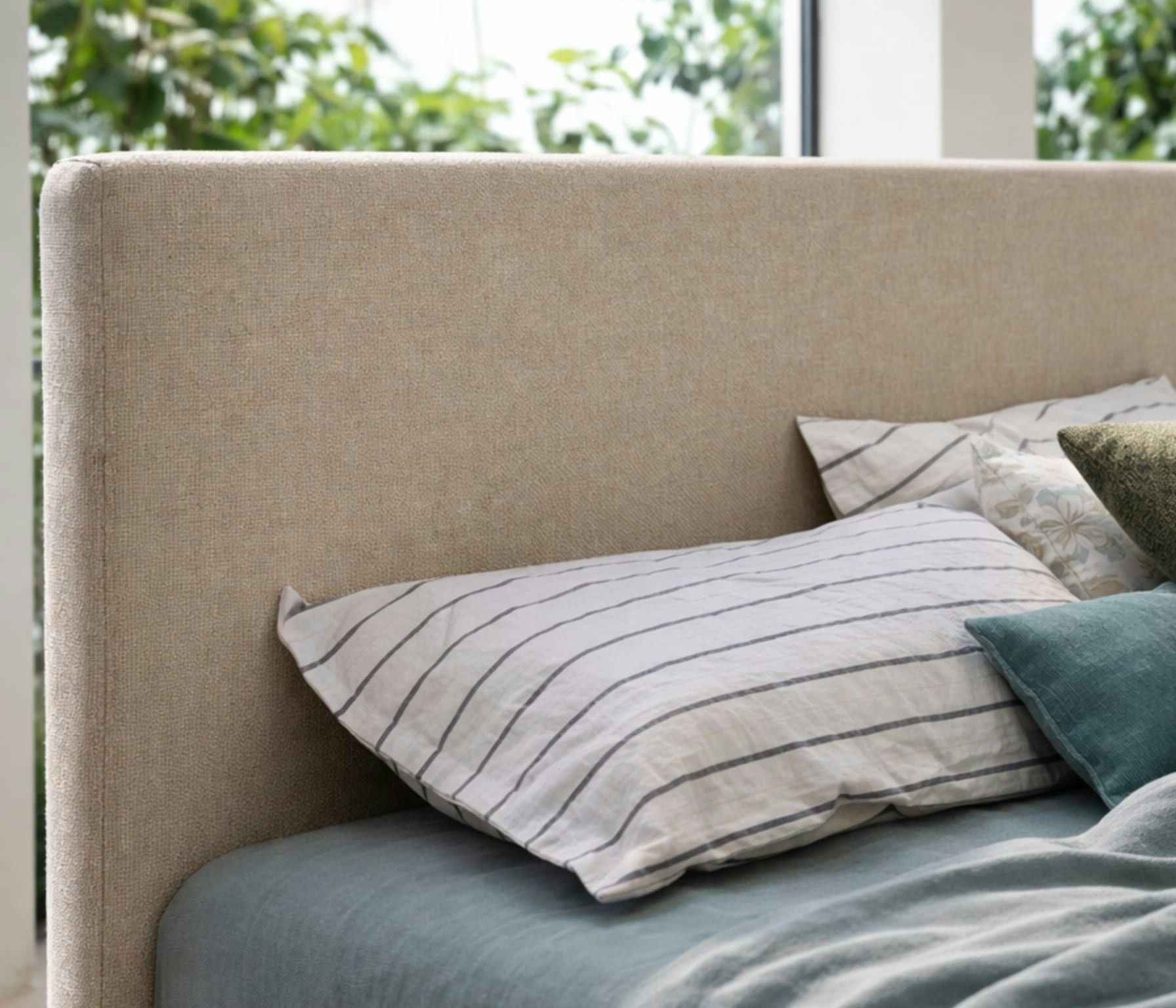 Letto con testiera beige in tessuto, cuscini con federe a righe e tinta unita, sfondo con finestra e piante verdi.