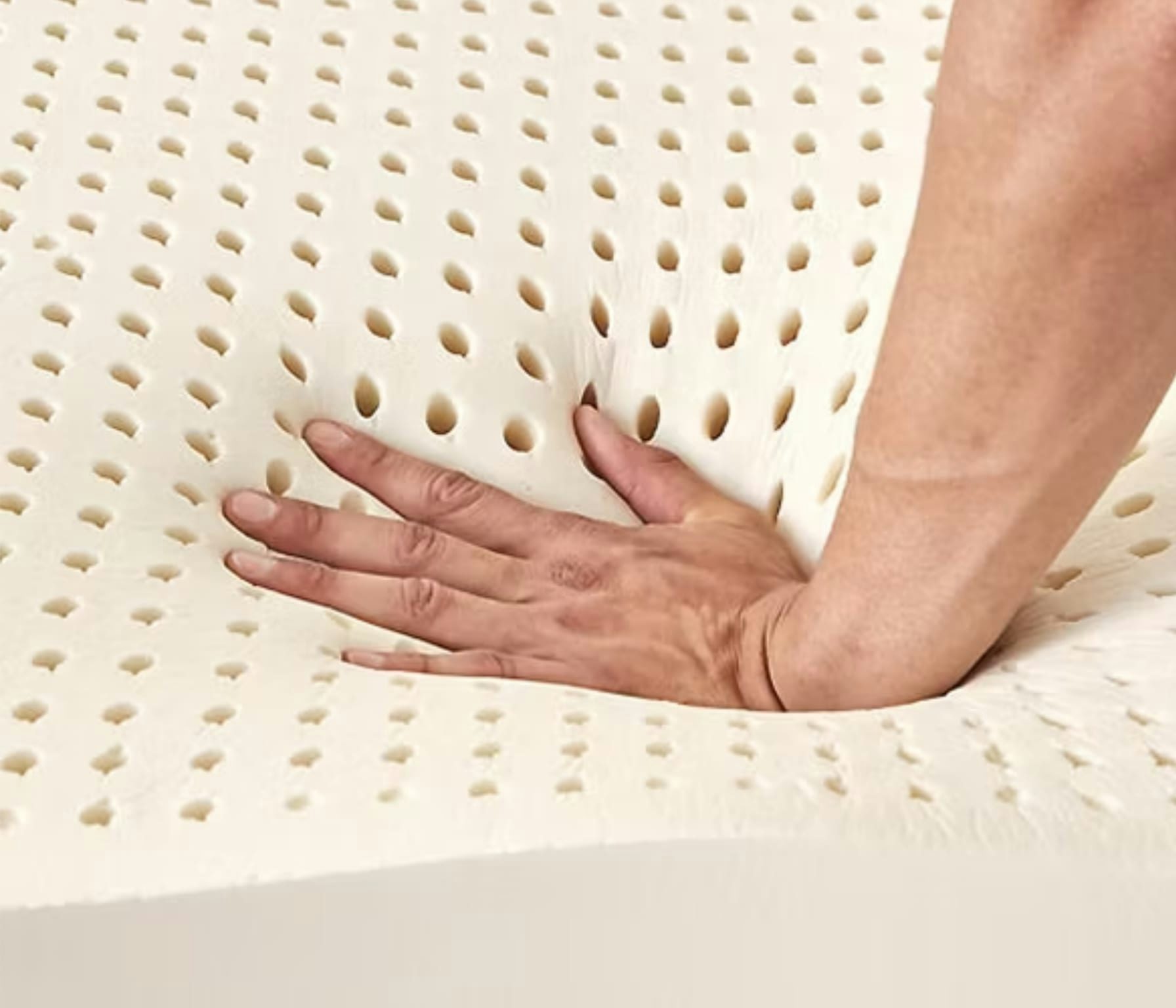 Mano che preme un materasso in lattice beige con fori traspiranti, evidenziando elasticità e morbidezza del materiale.