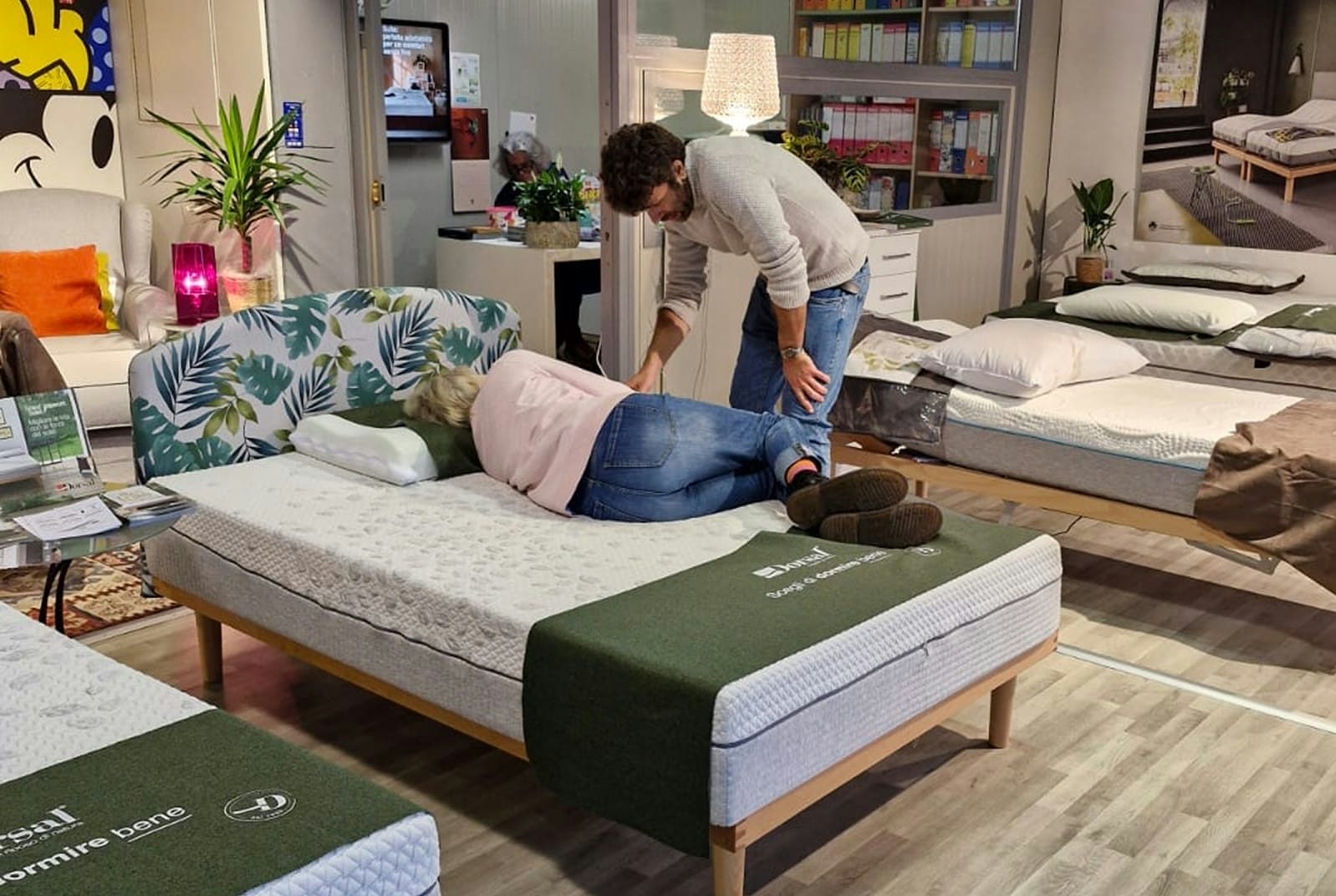 Donna sdraiata di lato su materasso con uomo che la osserva in negozio di arredamento per il sonno.