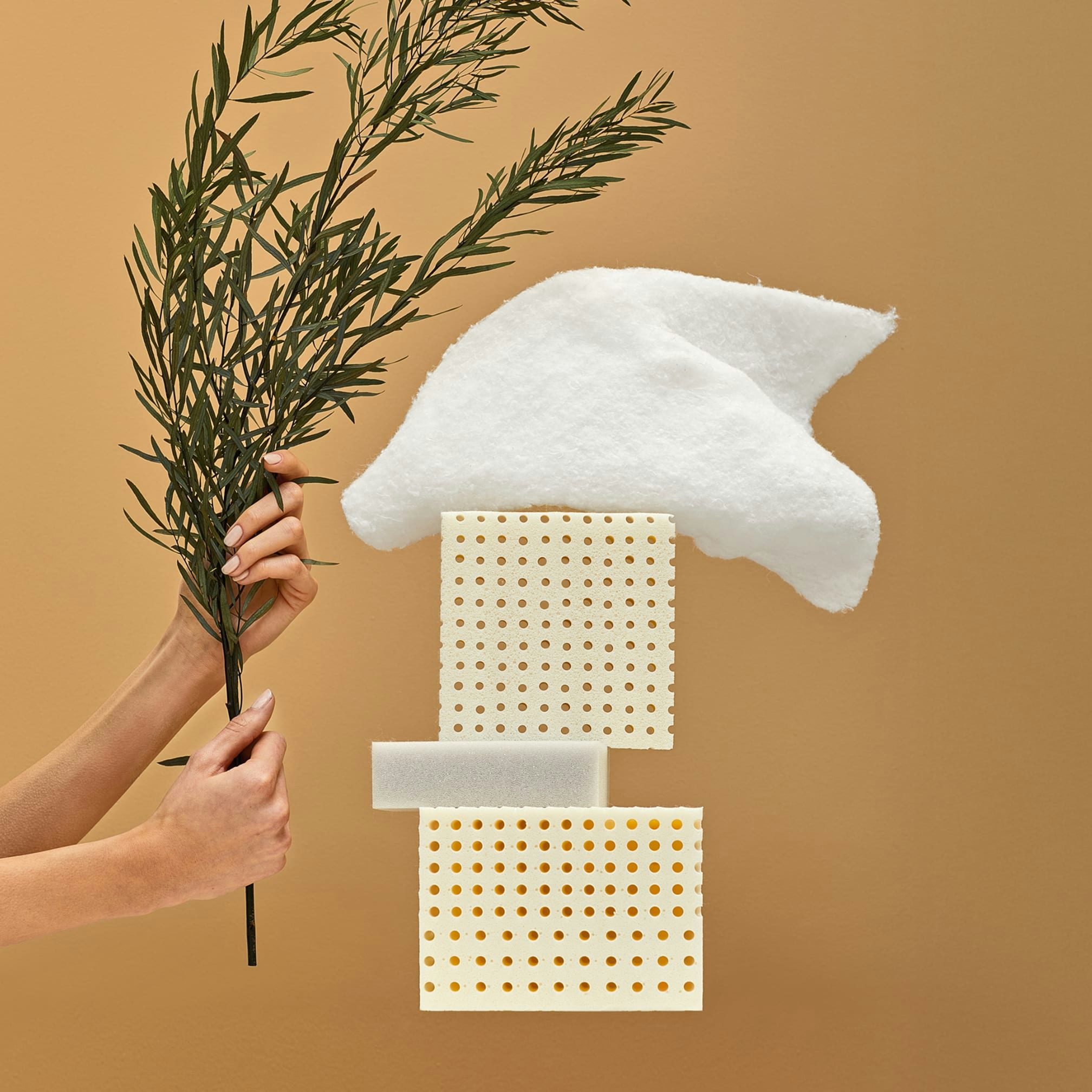 Mani che tengono un ramo verde accanto a tre blocchi di materiale e un pezzo di tessuto bianco su sfondo marrone chiaro.