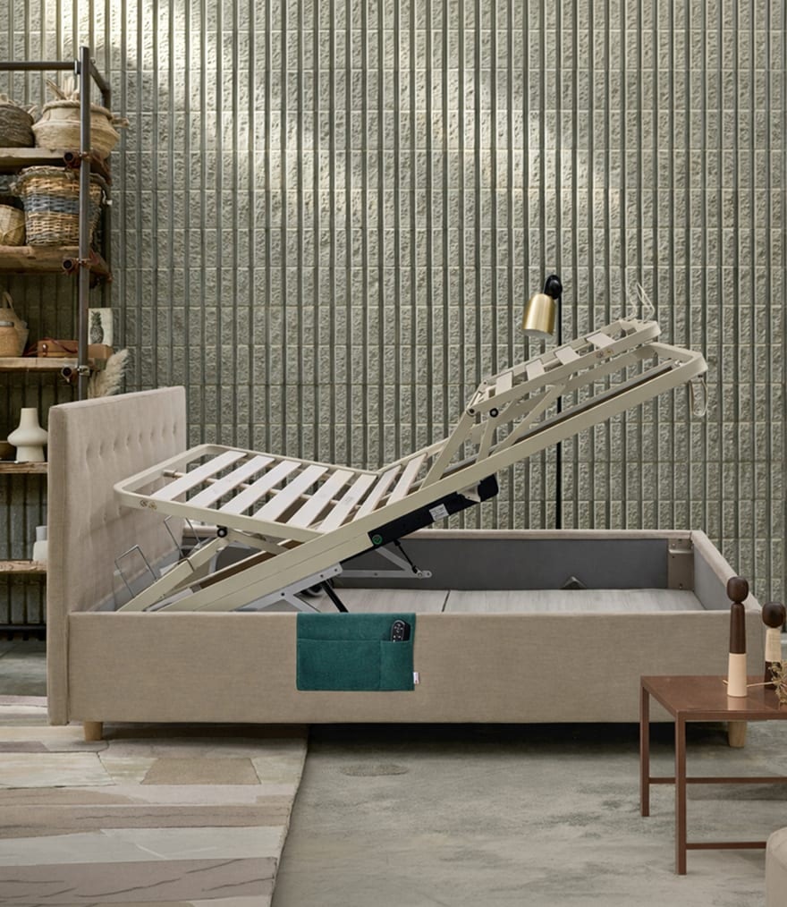 Letto matrimoniale beige con rete motorizzata sollevata, accanto scaffale in legno e tavolino marrone con decorazioni.