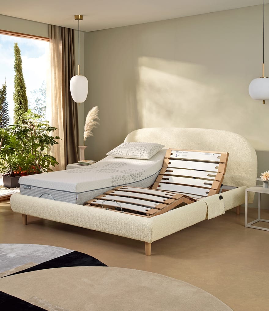 Letto matrimoniale regolabile in tessuto beige con materasso e cuscino bianco, piante vicino a finestra e lampade sospese.