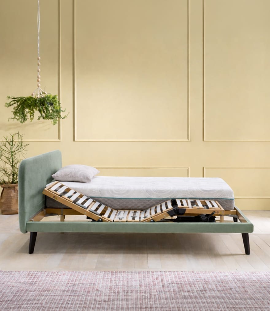 Letto con struttura regolabile in legno e imbottitura verde, materasso bianco e cuscino grigio, piante e parete beige.